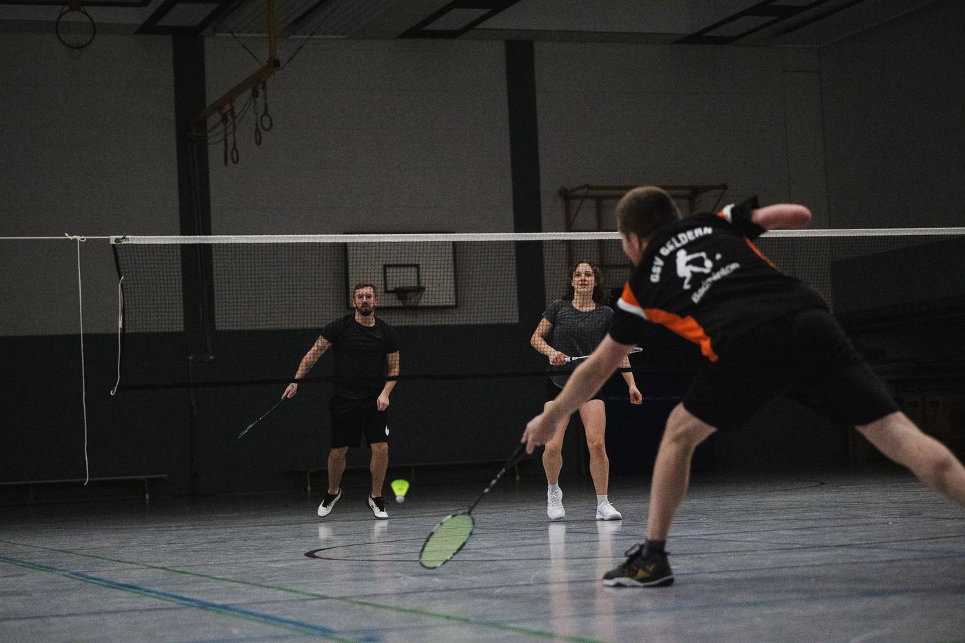 Doppel im Spiel: Ein Spieler im orange-schwarzen Trikot holt am Netz zum Schlag aus, drei Mitspielende stehen auf der anderen Netzseite bereit.