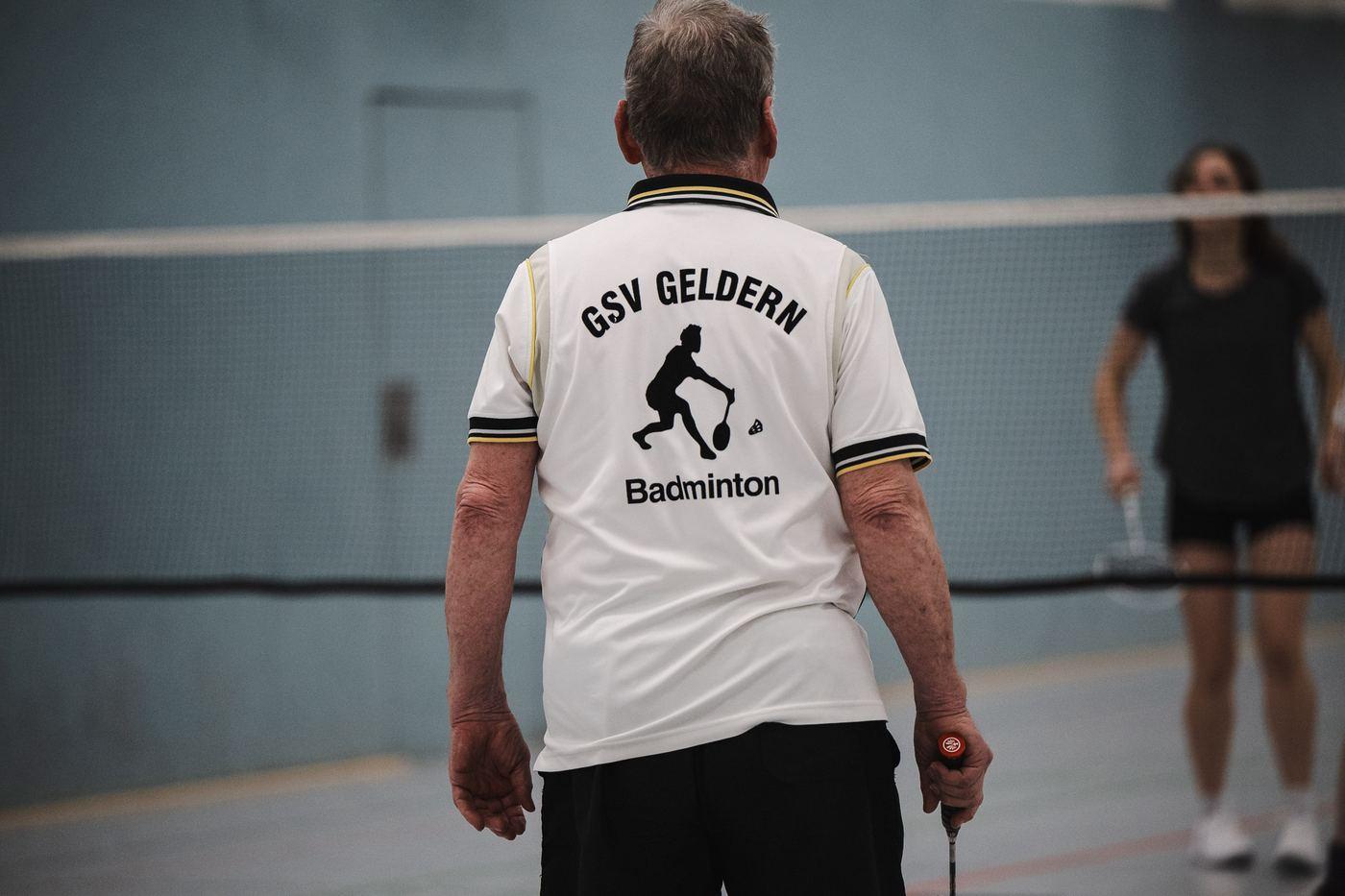 Ein Spieler im weißen Vereinsshirt mit dem Aufdruck „GSV Geldern Badminton“ steht mit dem Rücken zur Kamera am Netz, dahinter unscharf seine Gegnerin.