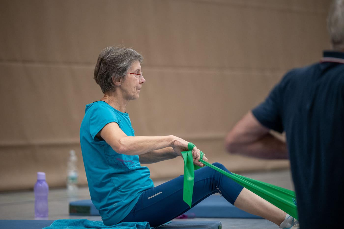 Eine Sportlerin sitzt auf der Matte und zieht das grüne Gymnastikband um ihren Fuß zu sich heran