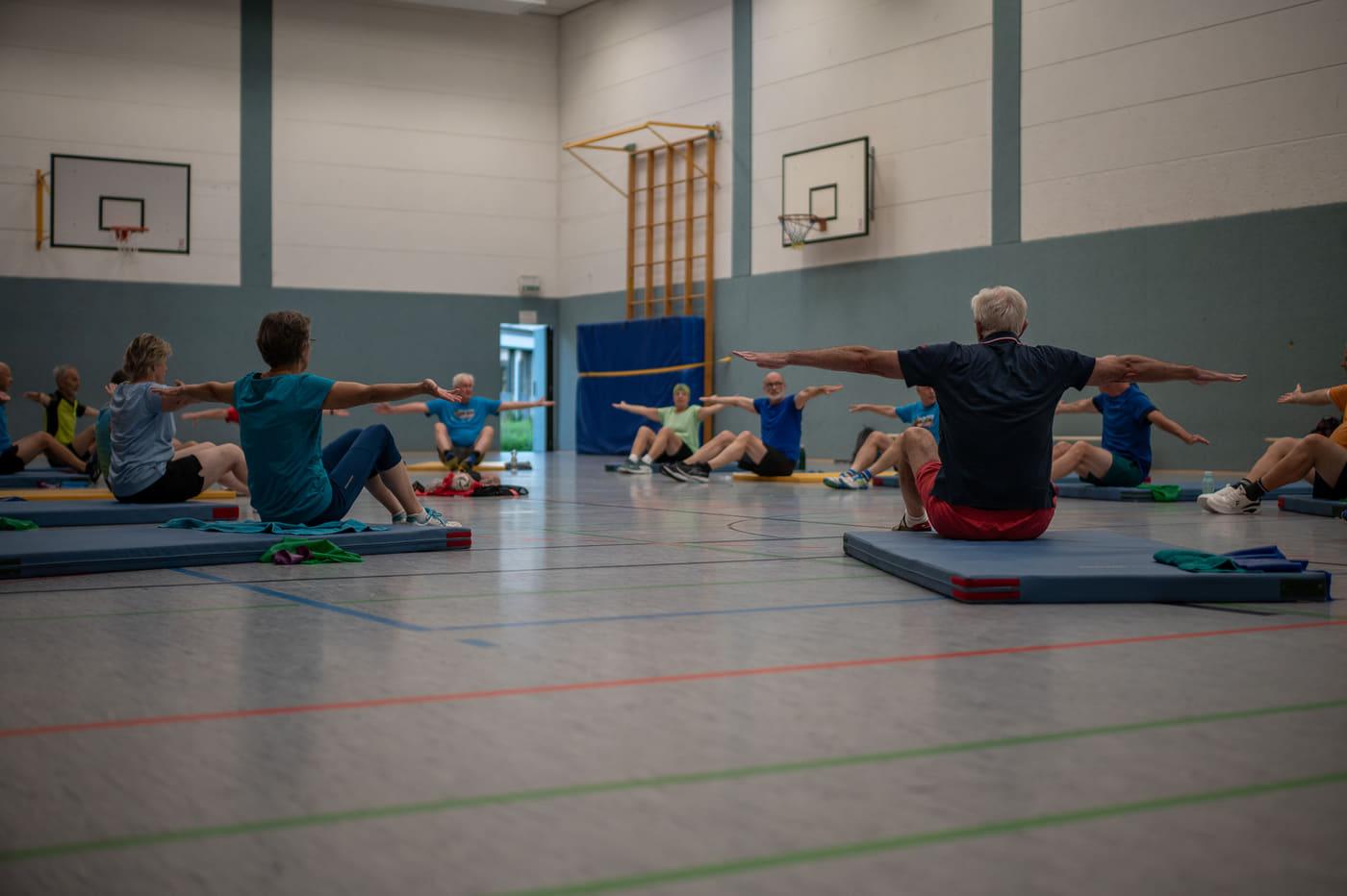 Die Gymnastikgruppe sitzt auf Matten in der Sporthalle und streckt die Arme zur Seite aus