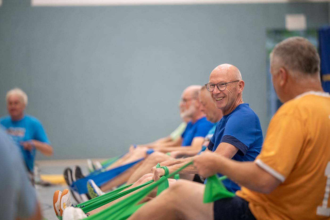 Mehrere Teilnehmende sitzen lachend nebeneinander und ziehen gemeinsam an grünen Gymnastikbändern