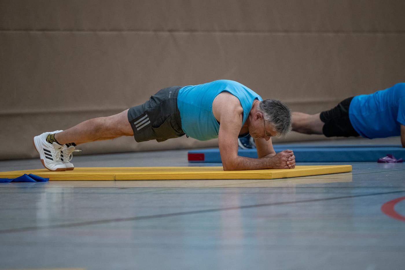 Ein Sportler hält den Unterarmstütz auf einer gelben Matte, neben ihm üben weitere in der Reihe