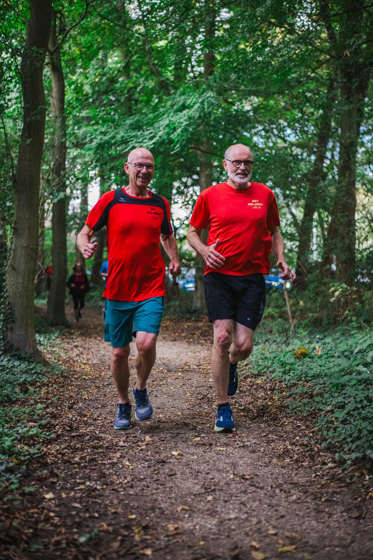 Zwei Läufer in roten Vereinsshirts laufen nebeneinander über den herbstlichen Waldweg