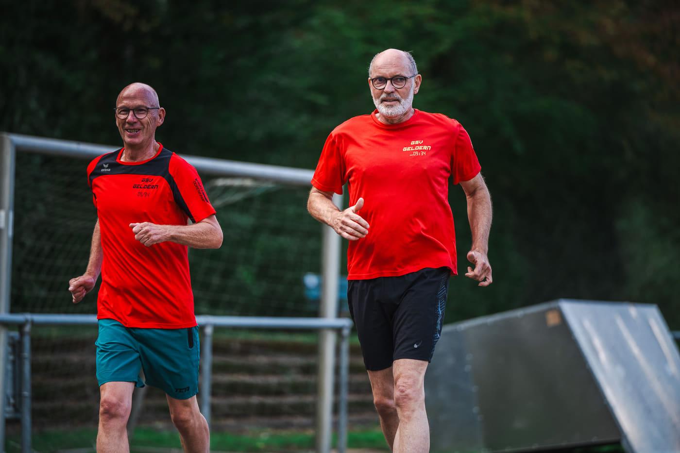 Zwei Läufer in roten Vereinsshirts traben nebeneinander am Sportplatz entlang