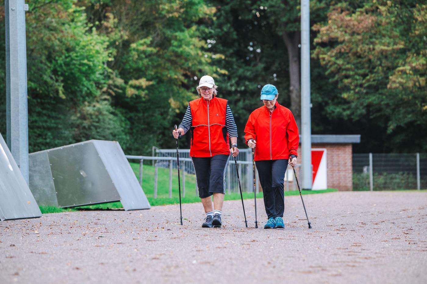 Zwei Nordic Walkerinnen in roten Jacken gehen mit Stöcken nebeneinander über den Weg am Stadion