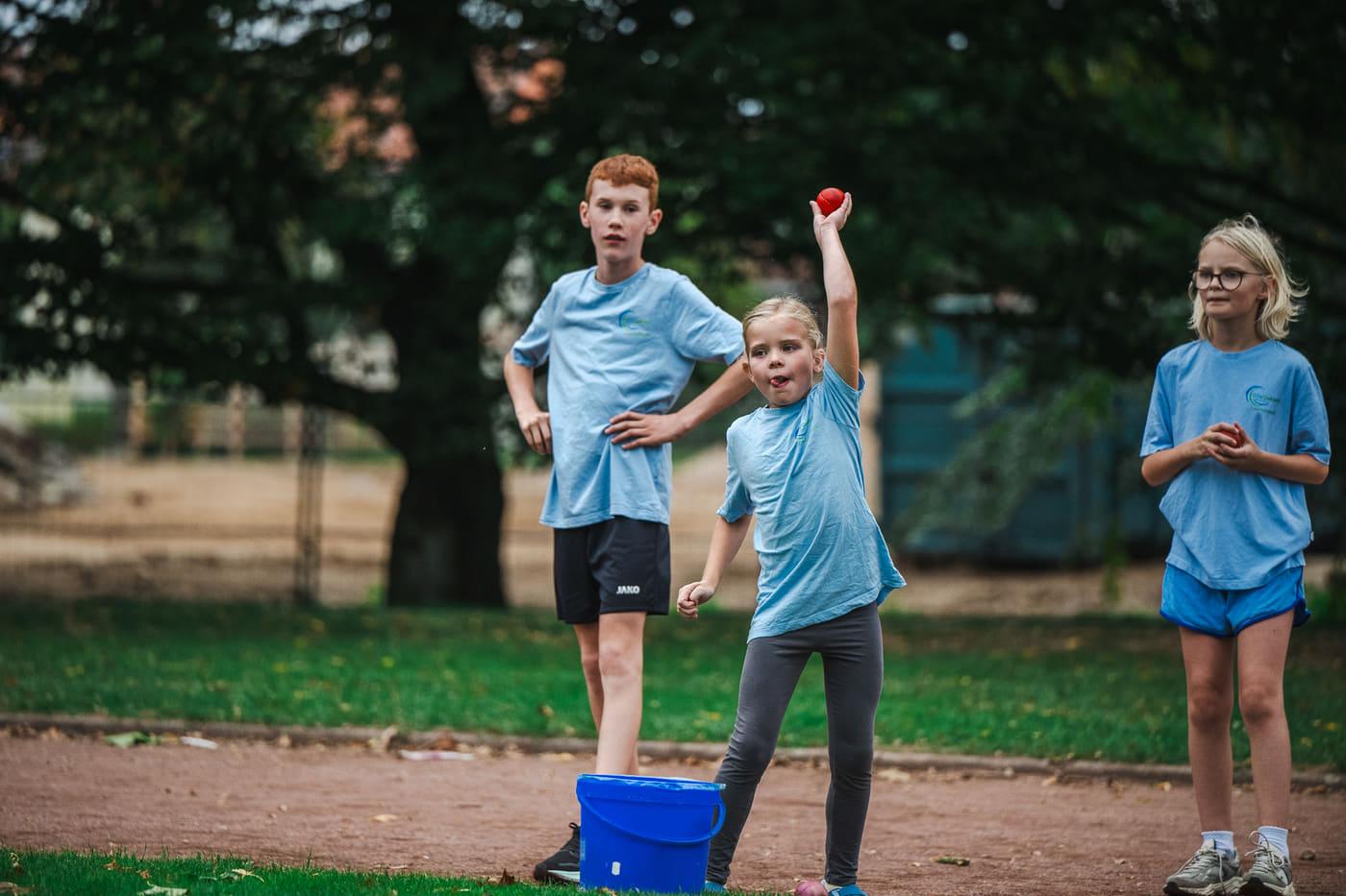 Mädchen wirft den roten Schlagball, neben ihr warten zwei Kinder am Eimer