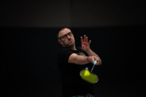 Nahaufnahme eines Spielers mit Brille, der den Blick intensiv auf den Federball richtet und gerade zum Schlag ausholt; der Hintergrund ist dunkel gehalten.