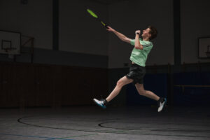 Ein Badmintonspieler in hellgrünem Trikot ist mitten im Sprung eingefangen, den Körper gestreckt, um den Federball in der Luft zu treffen.
