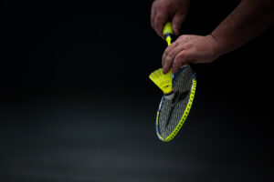 Nahaufnahme von Händen, die einen Badmintonschläger und einen gelben Federball halten, kurz vor dem Aufschlag vor einem komplett schwarzen Hintergrund.