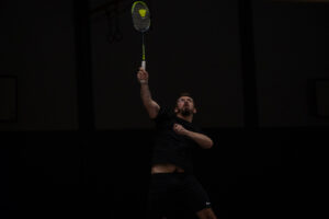 Ein Spieler springt mit dem Rücken zur Kamera hoch zum Schlag, während sein Doppelpartner zuschaut und ein gegnerischer Spieler auf der anderen Netzseite bereitsteht.