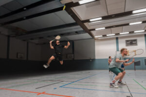 Ein Badmintonspieler in schwarzer Kleidung springt hoch in die Luft, um einen Schmetterschlag auszuführen, während ein Mitspieler in grüner Hose defensiv in der Hocke bereitsteht.