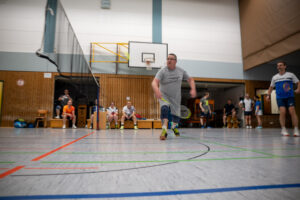 Ein Badmintonspieler in grauem T-Shirt macht einen weiten Ausfallschritt nach vorne ans Netz, um den Ball zu erreichen, während andere Spieler im Hintergrund in der Halle zusehen.